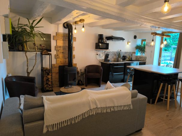 Vue d’ensemble d’un salon ouvert sur une cuisine moderne, avec poêle à bois, fauteuils en cuir et îlot central, dans une ambiance chaleureus