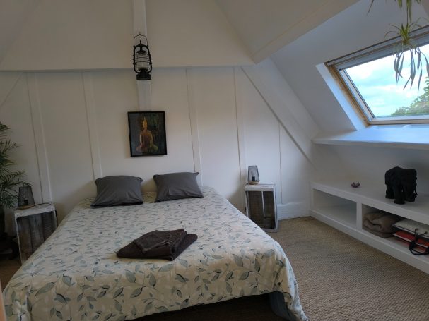 Chambre lumineuse à l’étage avec lit double, toit mansardé, décoration sobre et grande fenêtre de toit laissant entrer la lumière du ciel.