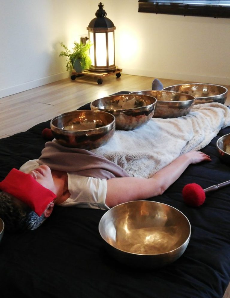 Soin sonore allongé avec bols tibétains Femme allongée dans une salle apaisante, éclairée à la lanterne, recevant un soin au bol tibétain avec plusieurs bols alignés sur le corps