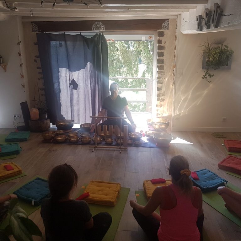 Un groupe de personnes assises en cercle sur des tapis, écoutant un praticien jouant des bols tibétains dans une salle lumineuse.
